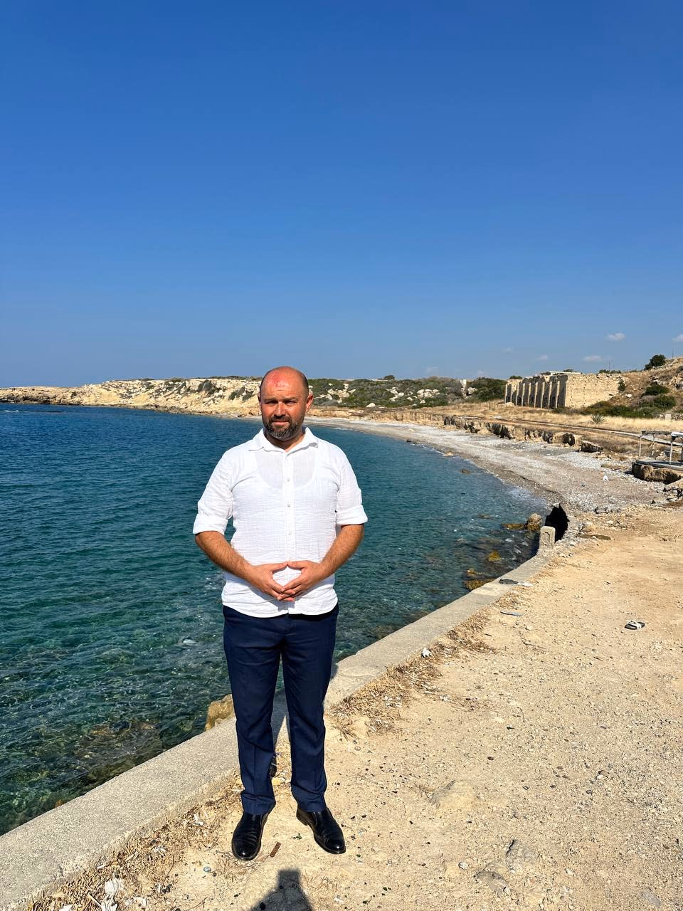 Doç. Dr. Muharrem Özdemir, Küçükerenköy sahilinde — arkada antik kalıntılar ve turkuaz koy
