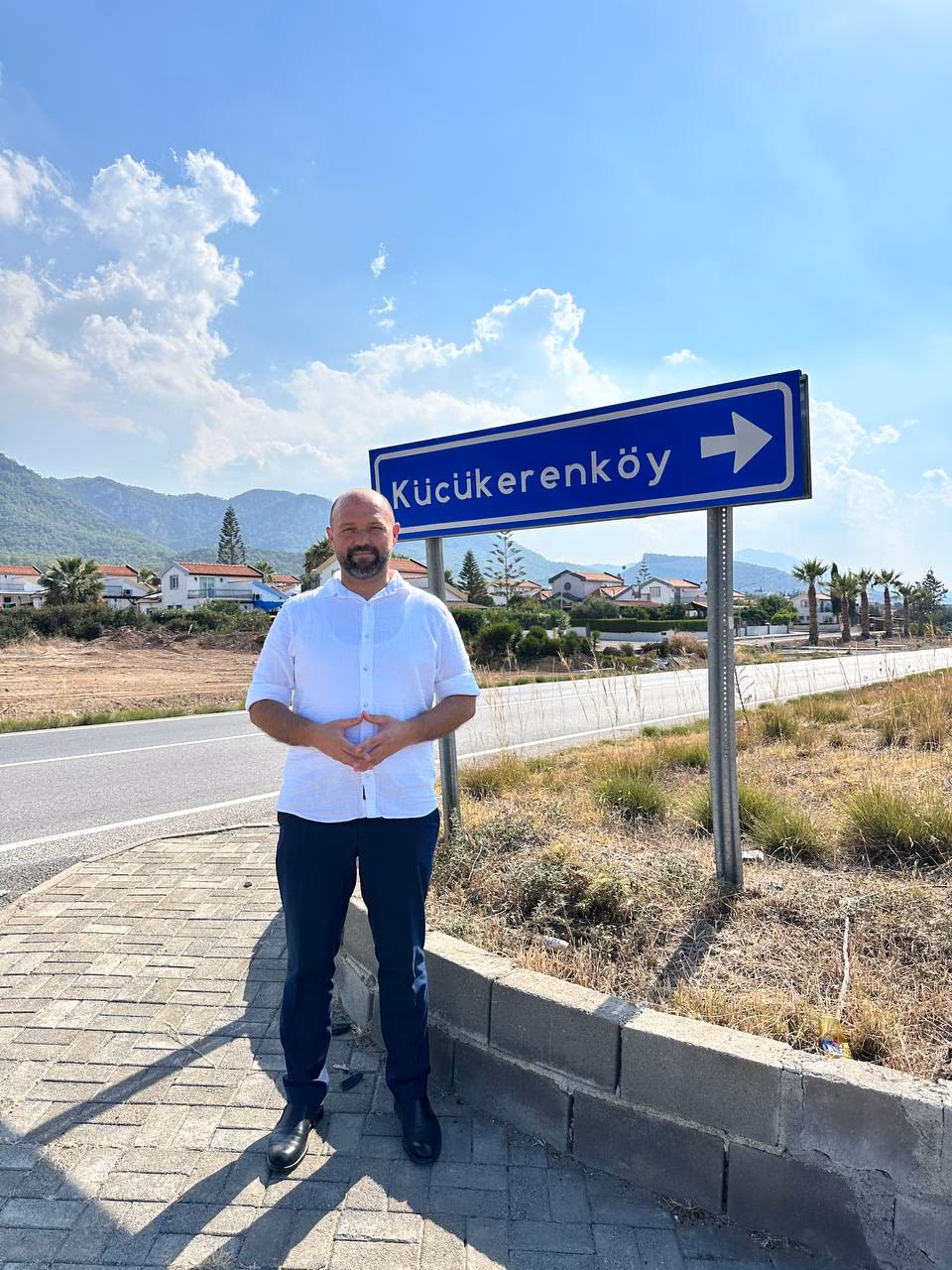 Doç. Dr. Muharrem Özdemir, Küçükerenköy tabelası önünde — arkada mahalle evleri ve Beşparmak Dağları