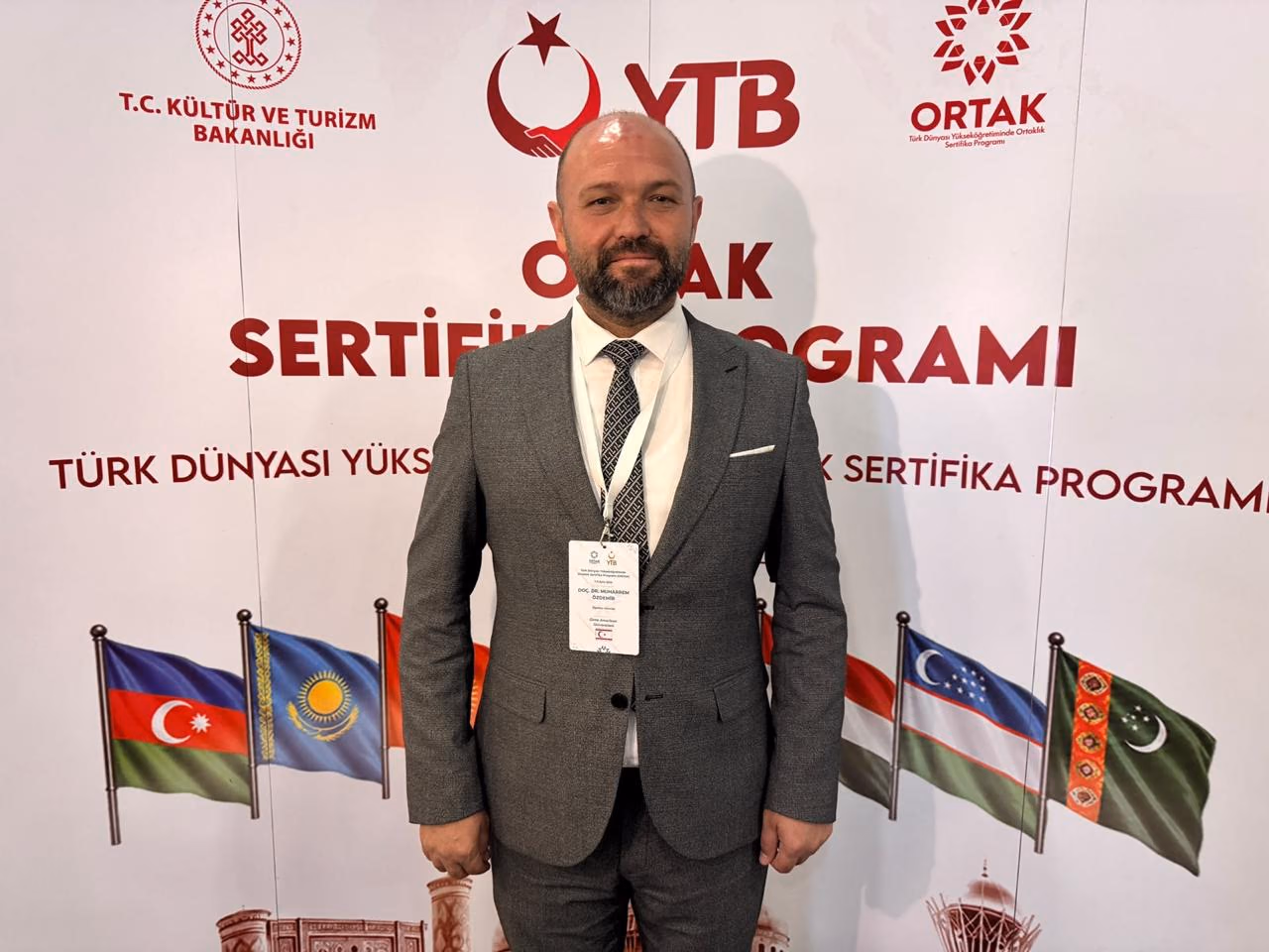 Doç. Dr. Muharrem Özdemir, YTB ORTAK Türk Dünyası Yükseköğretiminde Ortaklık Sertifika Programı afişi önünde — Türk dünyası bayrakları